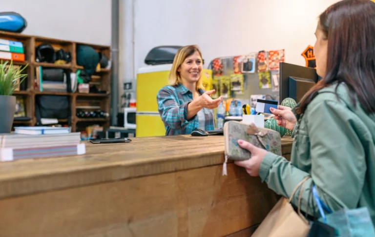 Mulher loira atrás de um balcão de madeira rústica estende a mão para receber um cartão de crédito azul de uma cliente em primeiro plano. O ambiente parece uma loja ou recepção de oficina, com prateleiras ao fundo.