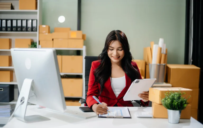 Mulher sorridente de blazer vermelho fazendo anotações em uma mesa, segurando um tablet na outra mão. Ao fundo, prateleiras com caixas de papelão.