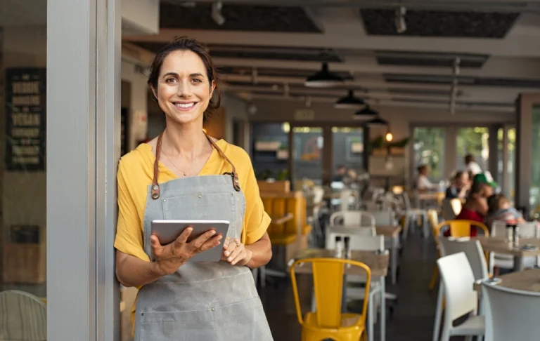 Mulher sorridente vestindo blusa amarela e avental cinza, em pé na entrada de um restaurante, segurando um tablet. Ao fundo, mesas, cadeiras vazias e luminárias pendentes criam um ambiente acolhedor.