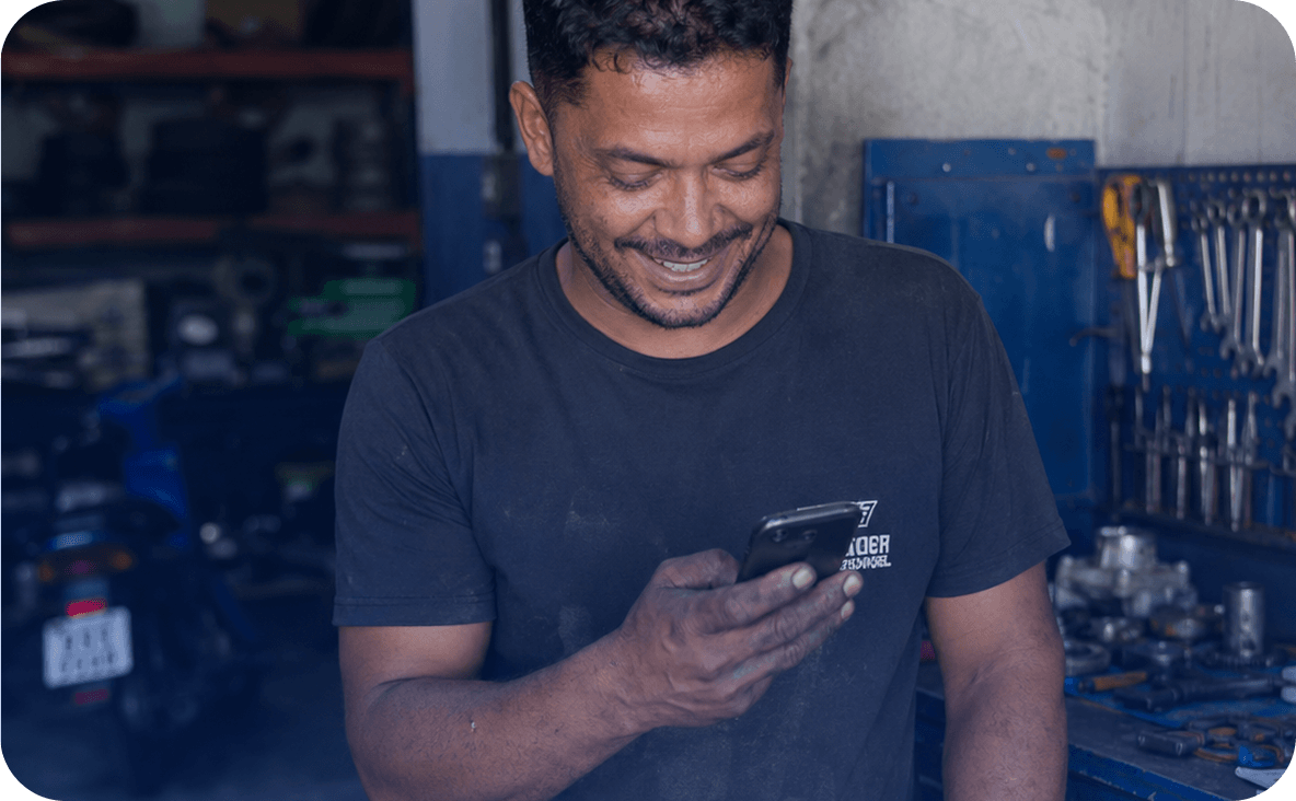 Homem sorridente com as roupas e as mãos sujas de graxa, segurando e olhando para a tela de um celular. O ambiente ao fundo é uma oficina mecânica, com um painel de ferramentas azul na parede e uma moto.