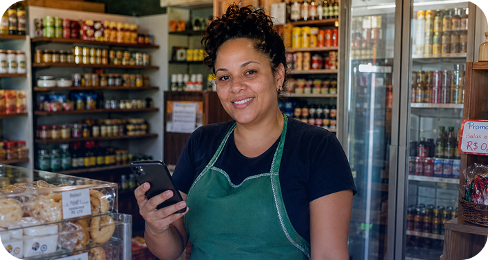 Mulher sorridente de cabelos cacheados presos, vestindo camiseta escura e avental verde. Ela segura um smartphone em uma mercearia, com prateleiras cheias de produtos ao fundo e uma estufa com pães à sua frente.
