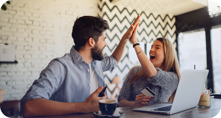 Homem e mulher sorrindo e batendo as mãos num gesto de comemoração (high five), sentados à mesa de uma cafeteria. A mulher segura um cartão de crédito e o homem um celular. Há um notebook branco aberto e xícaras de café na mesa.