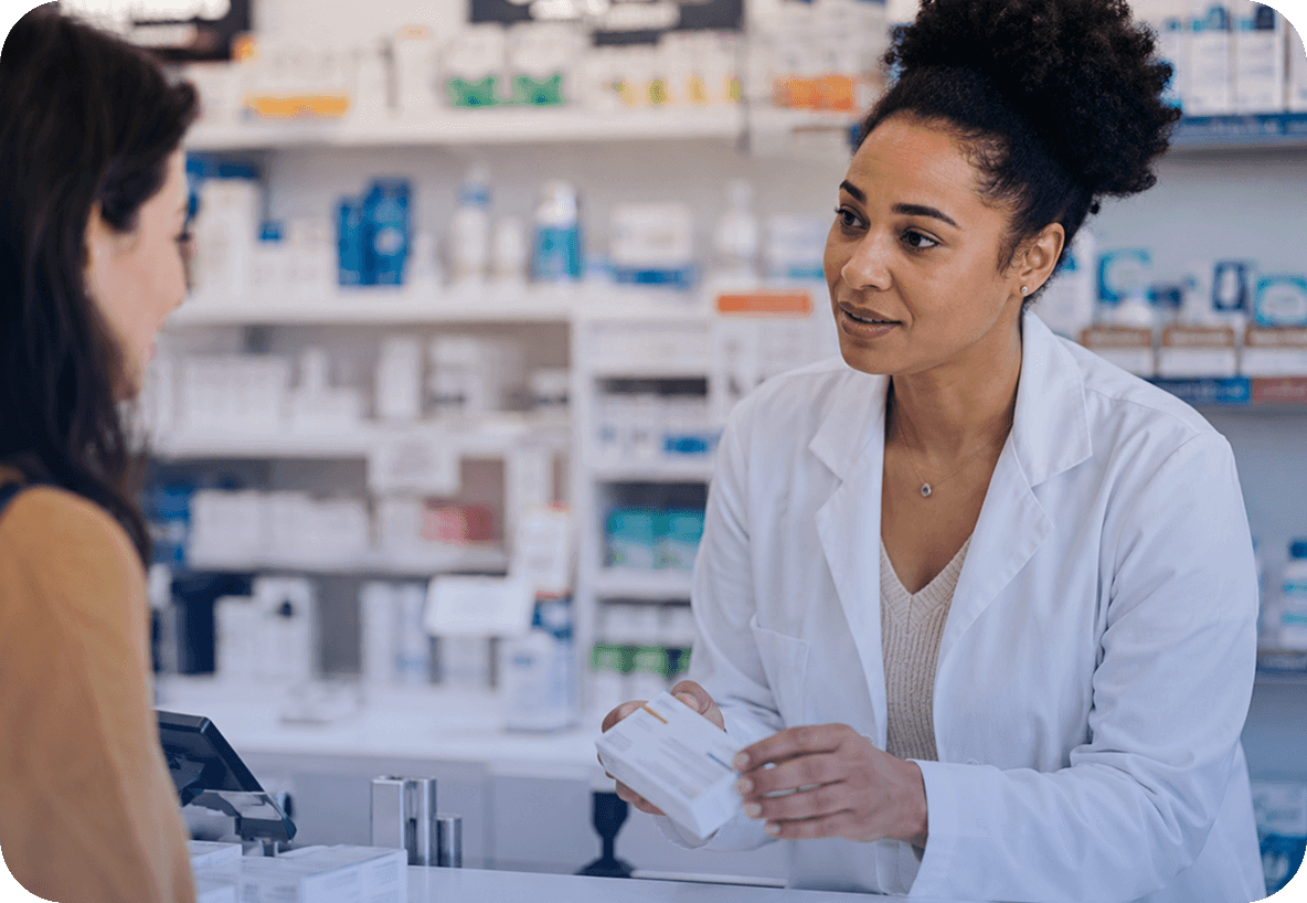 Mulher usando jaleco branco conversando com uma cliente no balcão de uma farmácia. Ela segura uma pequena caixa branca nas mãos. Ao fundo, prateleiras repletas de medicamentos