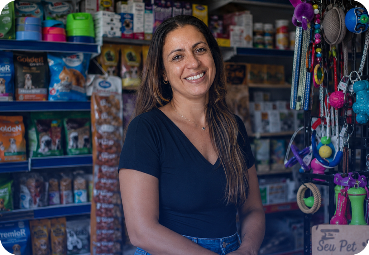 Mulher sorridente de cabelos longos e escuros, vestindo blusa preta, em pé no corredor de um pet shop. Ao fundo, prateleiras com pacotes de ração e brinquedos coloridos para animais pendurados.