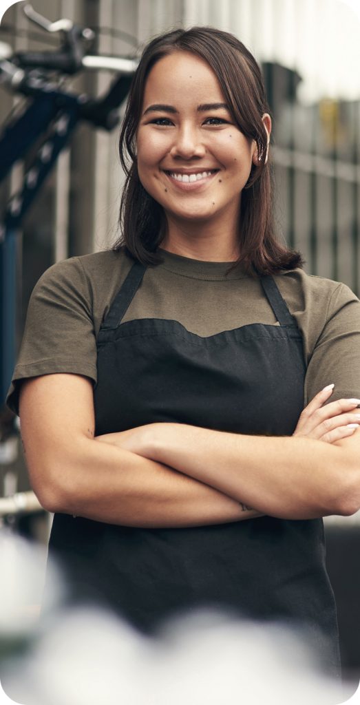 Mulher sorridente com cabelos escuros na altura dos ombros, vestindo camiseta verde-oliva e avental preto. Ela está de braços cruzados, posando em frente a uma estrutura metálica desfocada ao fundo.