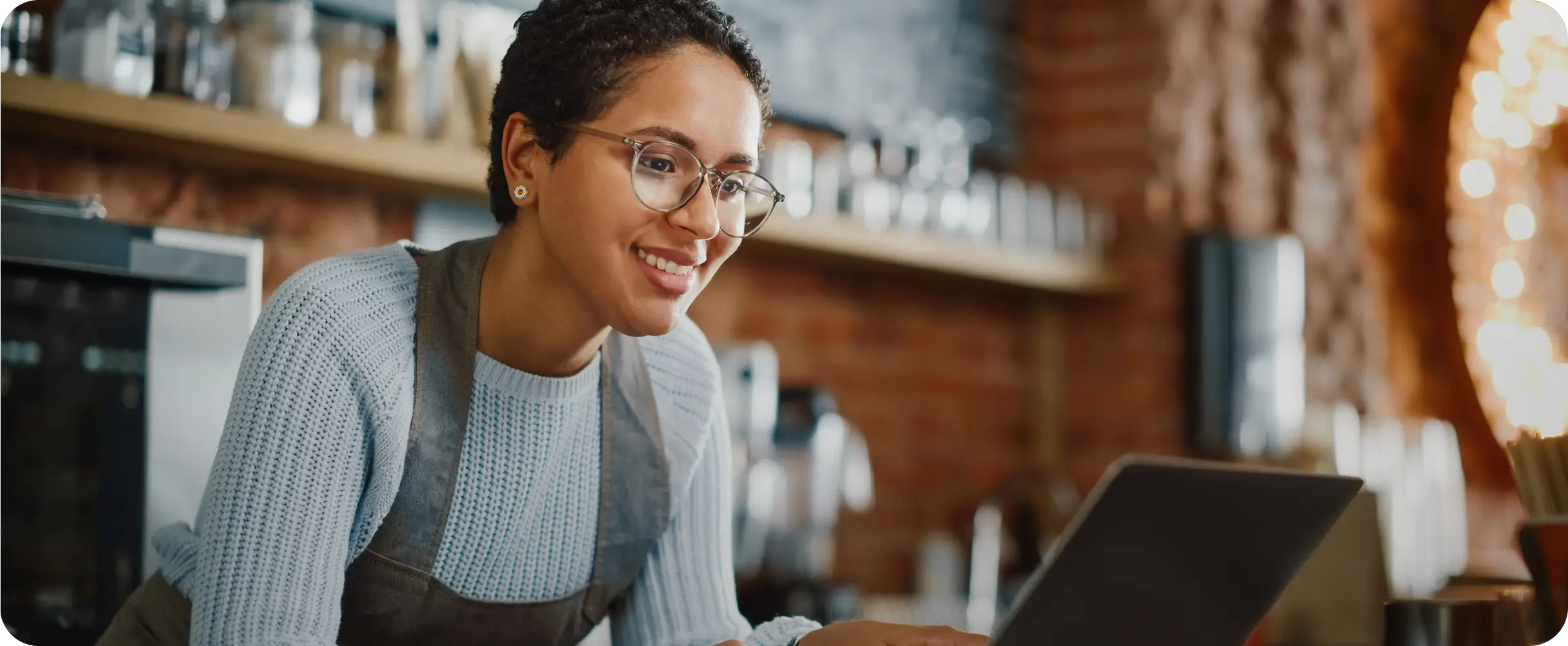 Mulher sorridente de óculos, vestindo suéter azul claro e avental cinza, inclinada sobre um balcão enquanto olha para a tela de um notebook. O fundo desfocado mostra o interior de um comércio com iluminação quente