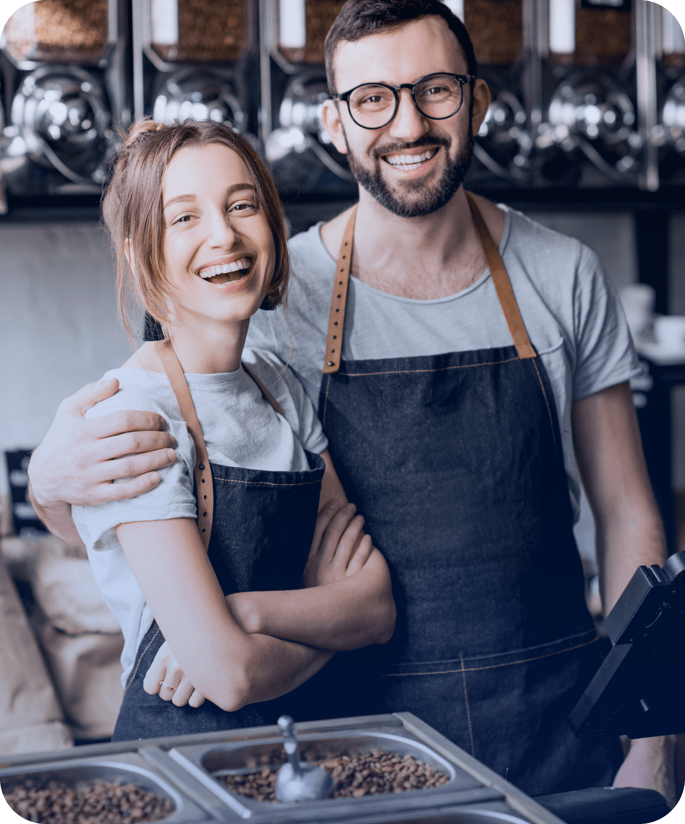 Homem com óculos e mulher, ambos sorridentes e usando aventais escuros, abraçados atrás de um balcão metálico contendo grãos de café. Ao fundo, paredes escuras com grandes dispensadores cheios de grãos de café.