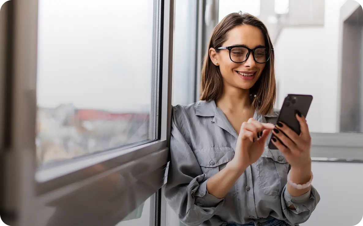 Mulher sorridente usando óculos de grau e camisa cinza, encostada perto de uma janela enquanto digita em um smartphone