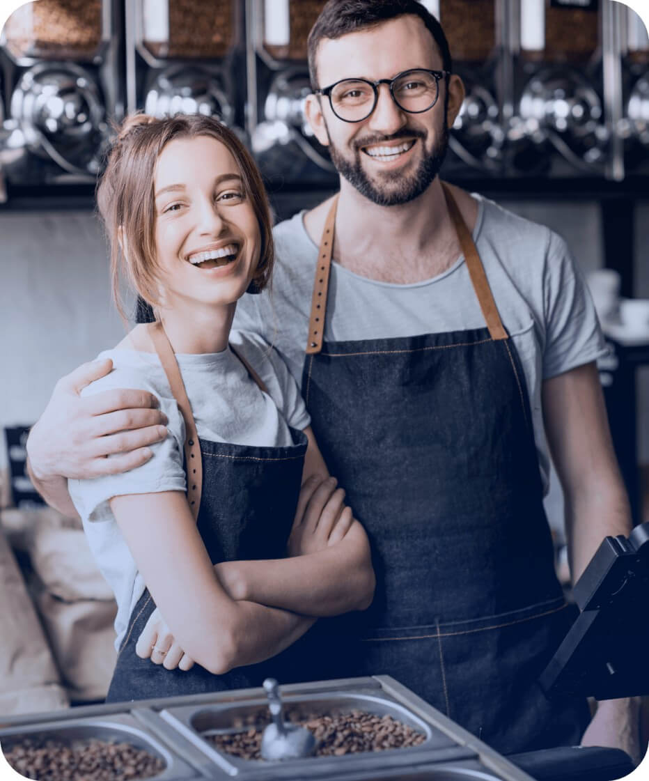 Homem com óculos e mulher, ambos sorridentes e usando aventais escuros, abraçados atrás de um balcão metálico contendo grãos de café. Ao fundo, paredes escuras com grandes dispensadores cheios de grãos de café.